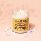 Glass jar candle with lit wick, featuring text "Two Hearts, One Mortgage, Home".