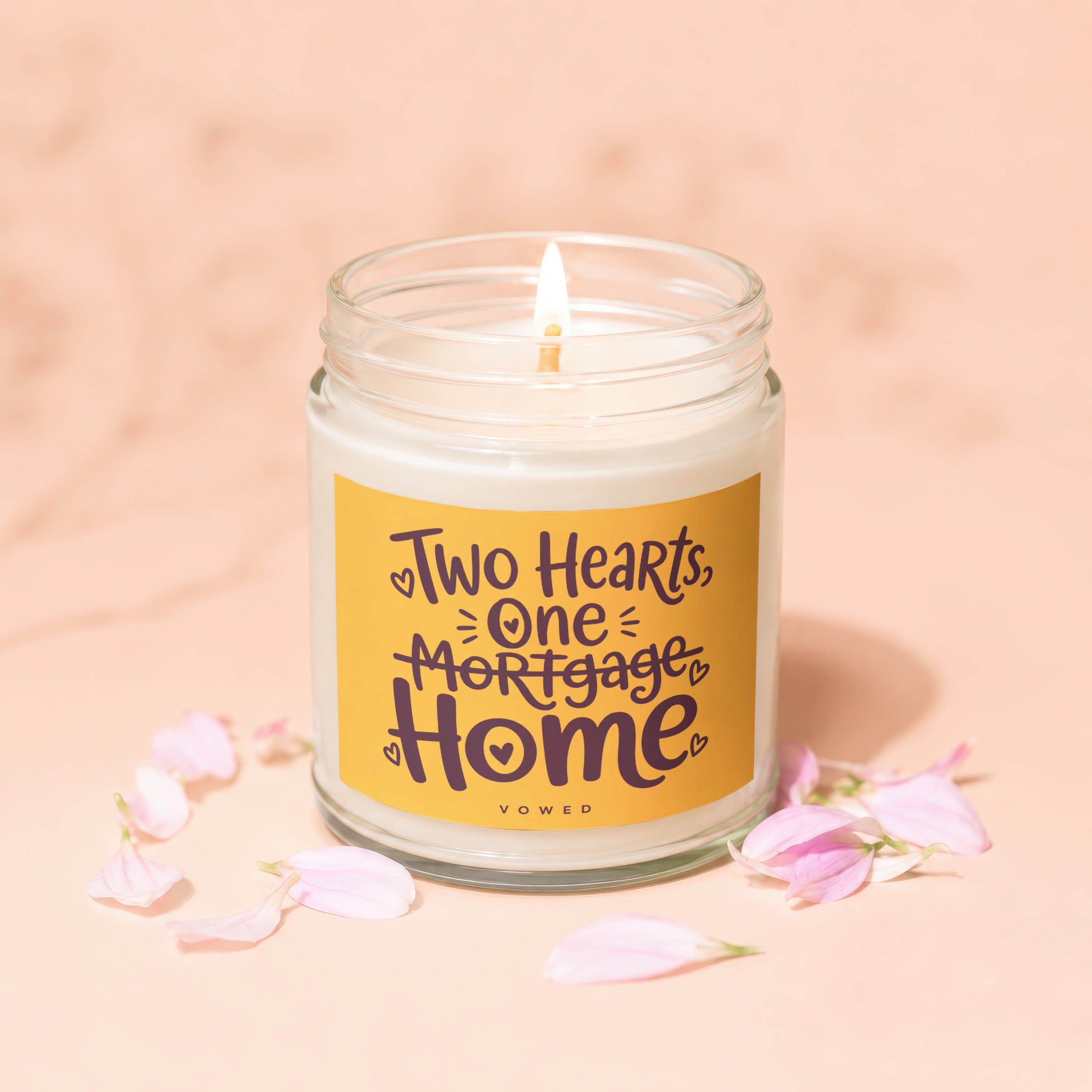 Glass jar candle with lit wick, featuring text "Two Hearts, One Mortgage, Home".