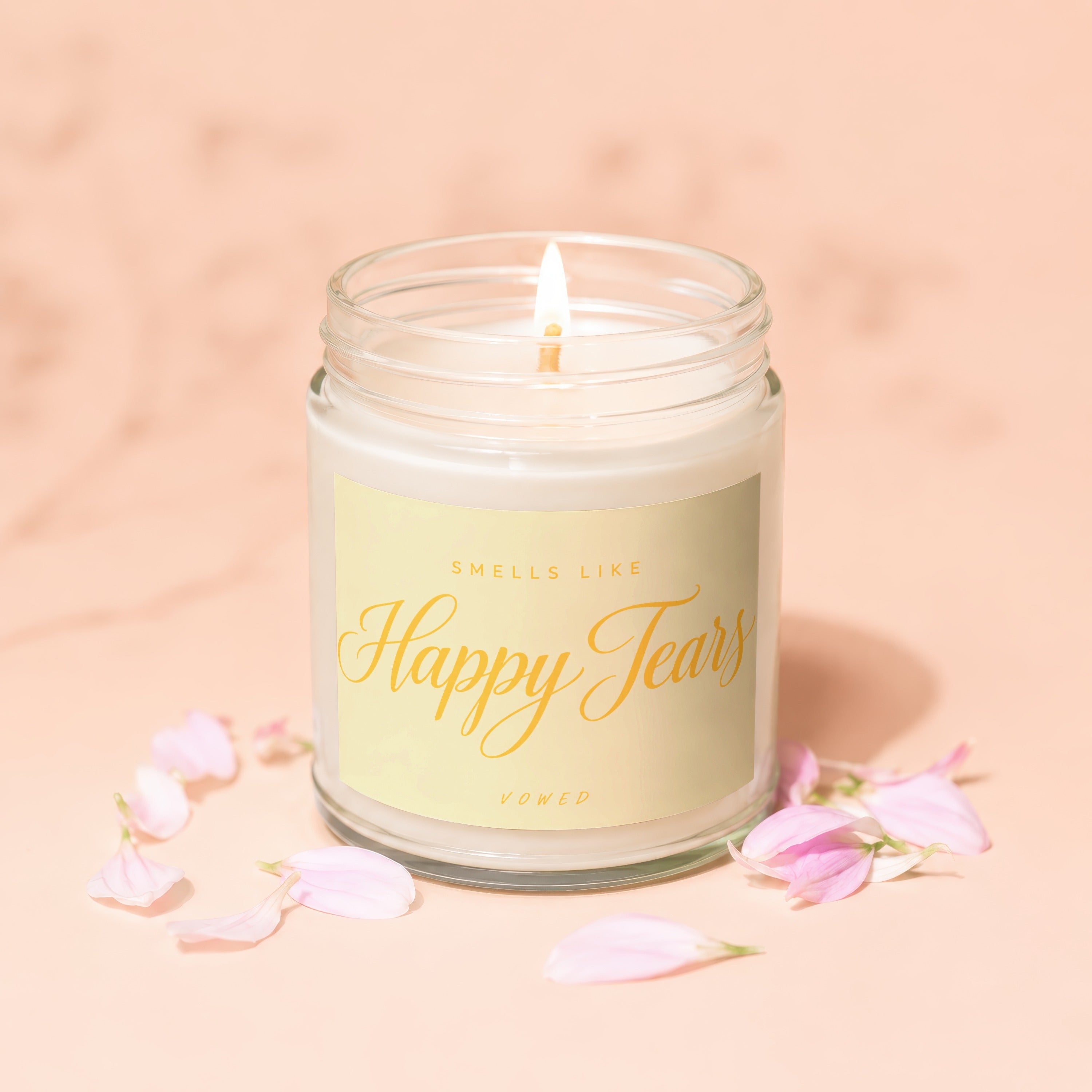 Lit candle in glass jar with "Smells Like Happy Tears" label surrounded by delicate petals.