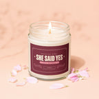 Candle with "She Said Yes" label, lit wick, and pink petals on peach background.