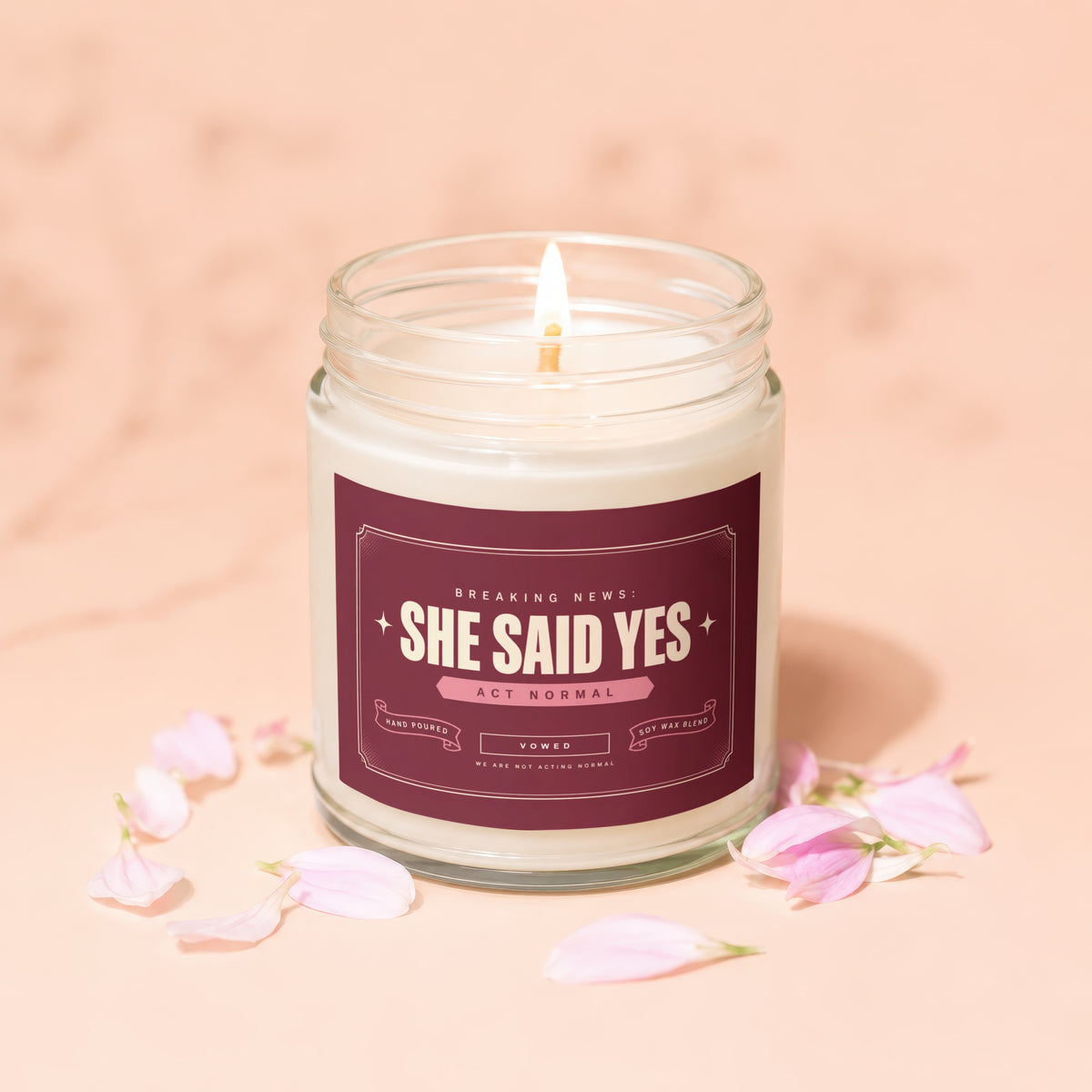 Candle with "She Said Yes" label, lit wick, and pink petals on peach background.