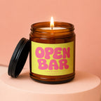 Amber jar candle with a lit wick and yellow label reading "OPEN BAR LOADING... VOWED".