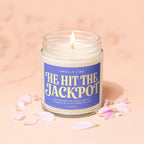 Lit candle in jar, reads "He Hit The Jackpot," surrounded by pink petals.