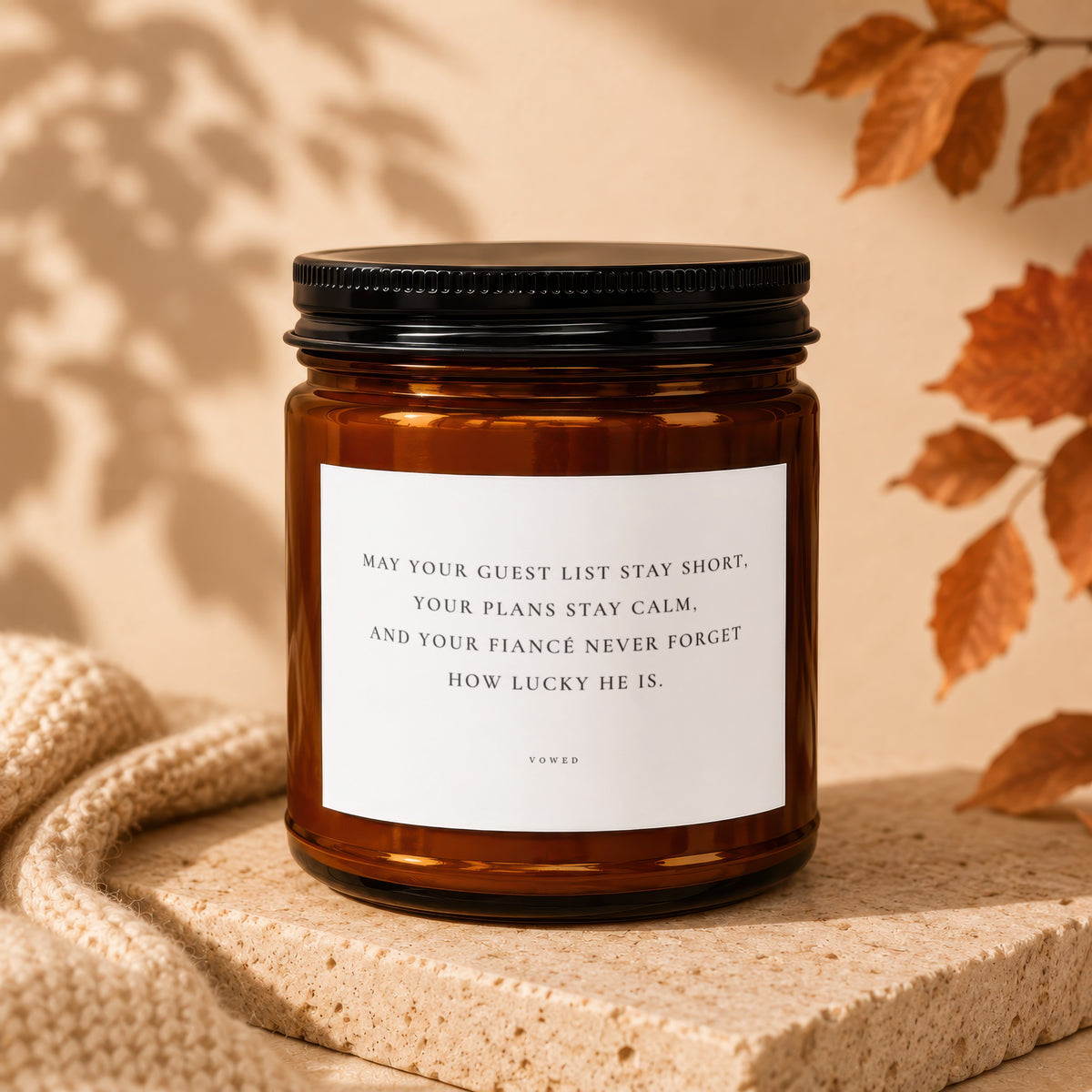 Amber glass candle with black lid and label reading: "May your guest list stay short, your plans stay calm, and your fiancé never forget how lucky he is. VOWED"