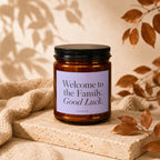 Amber candle with black lid and lavender label saying "Welcome to the Family. Good Luck."