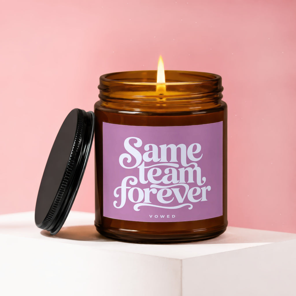Amber jar candle with lid askew, lit candle, purple label reads "Same team forever," "vowed".