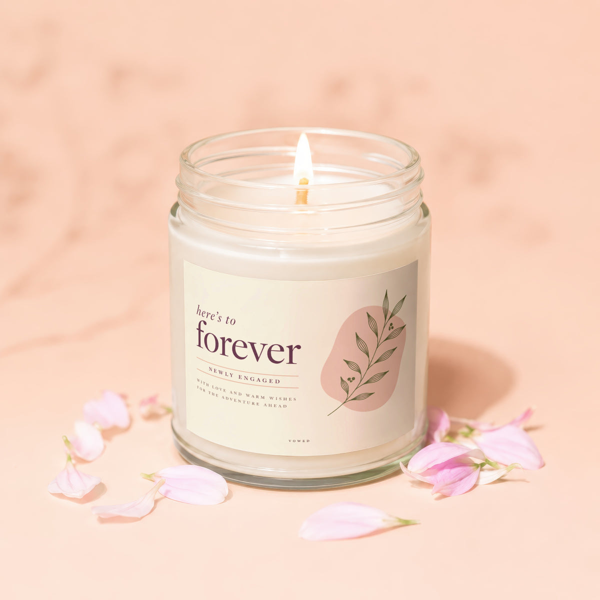 A lit candle in a glass jar with a label reading here's to forever, surrounded by pink petals.
