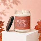 Glass candle with lit wick, "Engaged & thrilled vowed" text on label, autumn leaves.