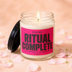 Glass candle with pink label reading "STATUS: RITUAL COMPLETE" surrounded by pink petals.