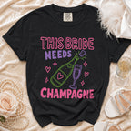 Black t-shirt with "This Bride Needs Champagne" text and champagne bottle graphic.