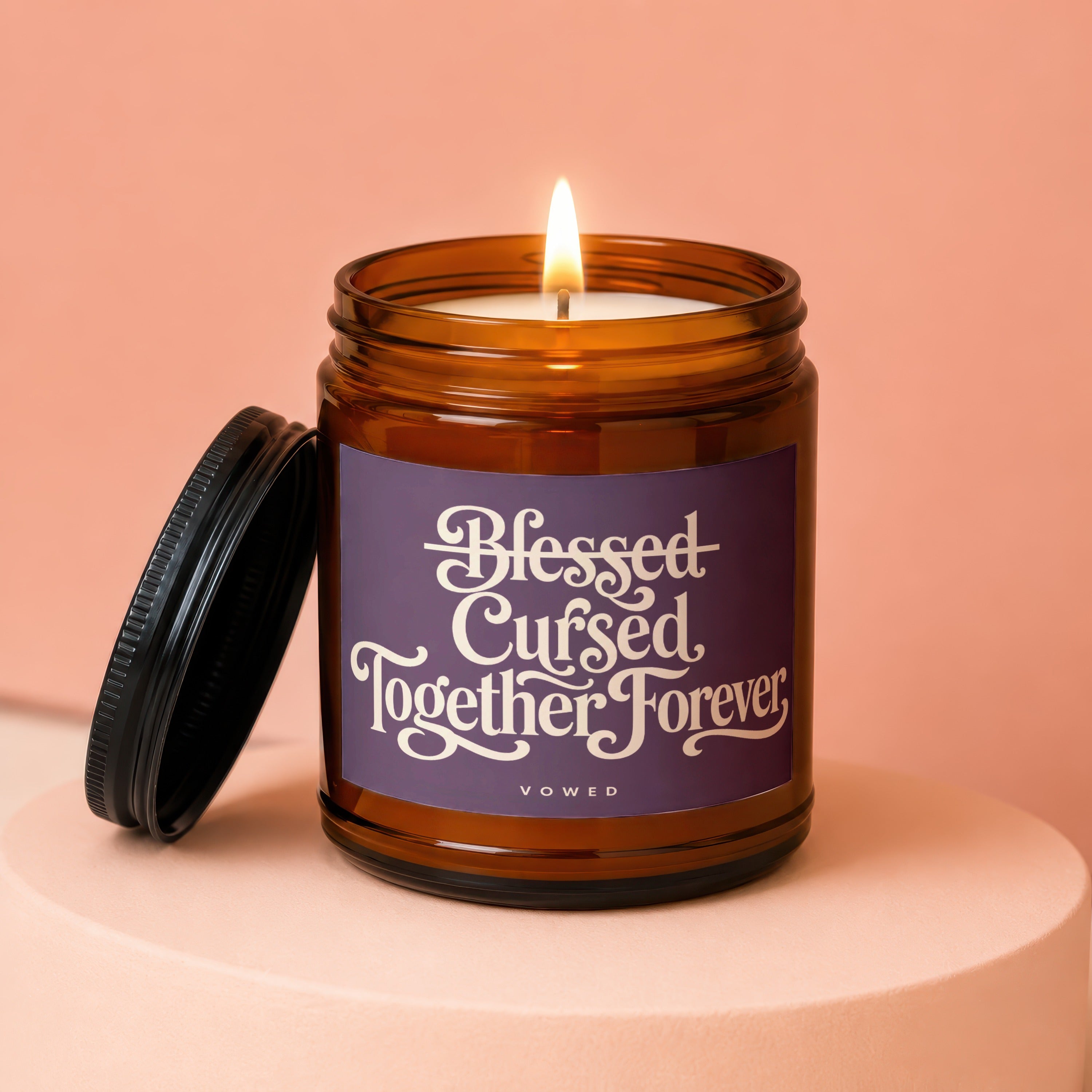 Amber jar candle with lit wick, purple label reads "Blessed Cursed Together Forever, VOWED".