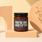 Amber jar candle with purple label reading "You're One Lucky B*tch".