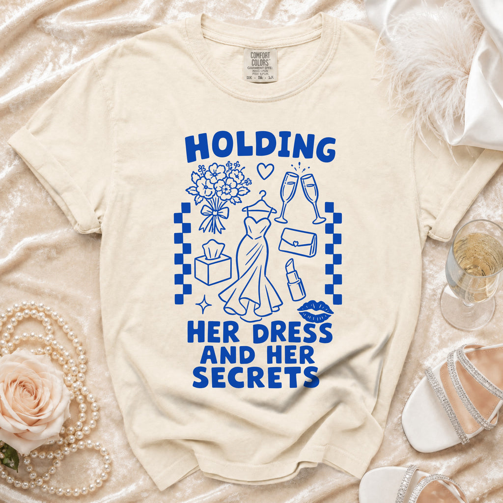Ecru t-shirt with blue text "HOLDING HER DRESS AND HER SECRETS" and graphics.