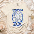 Ecru t-shirt with blue text "HOLDING HER DRESS AND HER SECRETS" and graphics.