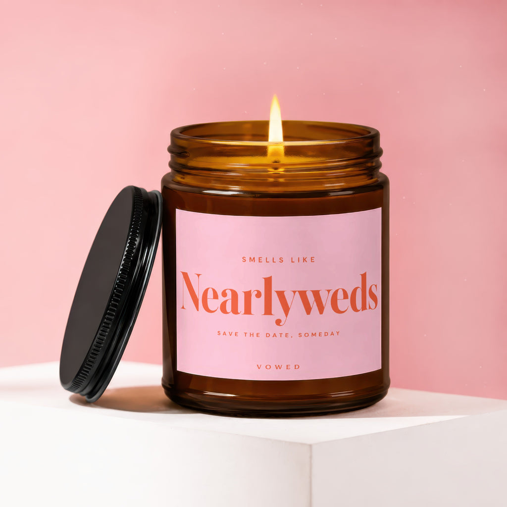 A lit amber jar candle with a pink label that reads, "Smells Like Nearlyweds Save the Date, Someday VOWED."
