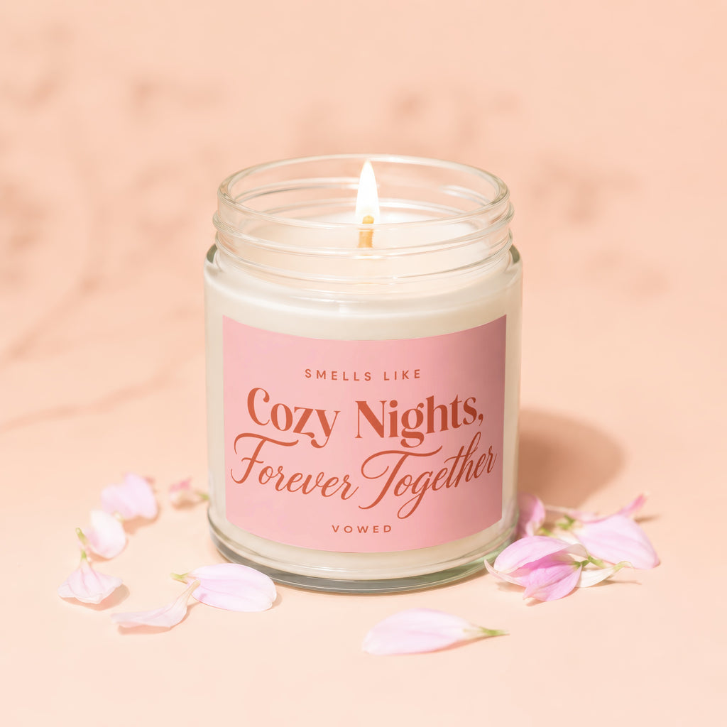Glass jar candle with lit wick and pink label reading "Smells like Cozy Nights, Forever Together"