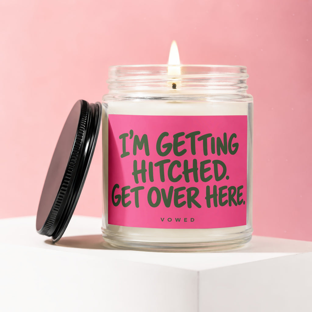 Glass candle with lit wick and black lid. Pink label reads "I'M GETTING HITCHED. GET OVER HERE."