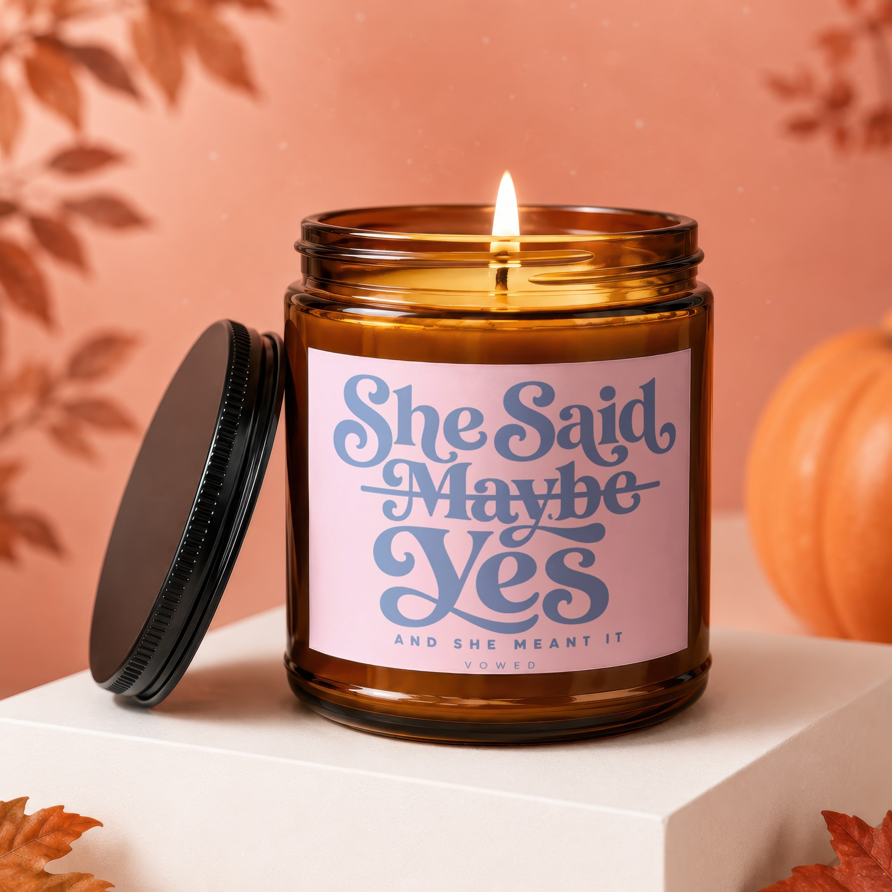 Amber jar candle with lit flame, pink label reads "She Said Maybe Yes"