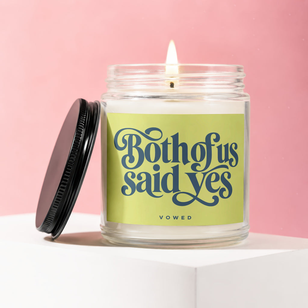 Glass candle with lit wick and black lid, labeled "Both of us said yes."
