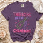 Purple t-shirt with "This Bride Needs Champagne" text and champagne bottle graphic.