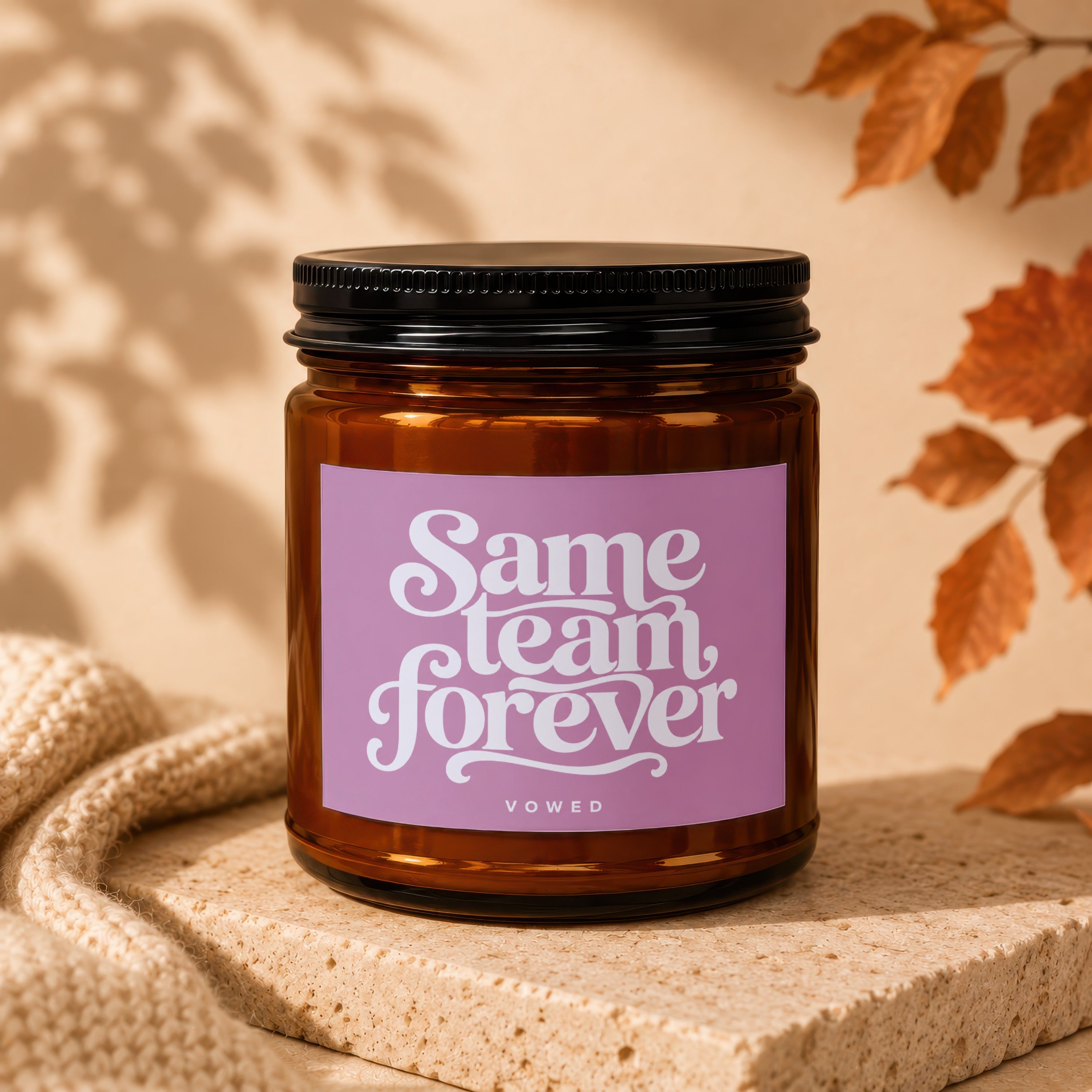 Amber jar candle with "Same team forever" text on a purple label, on a stone pedestal.