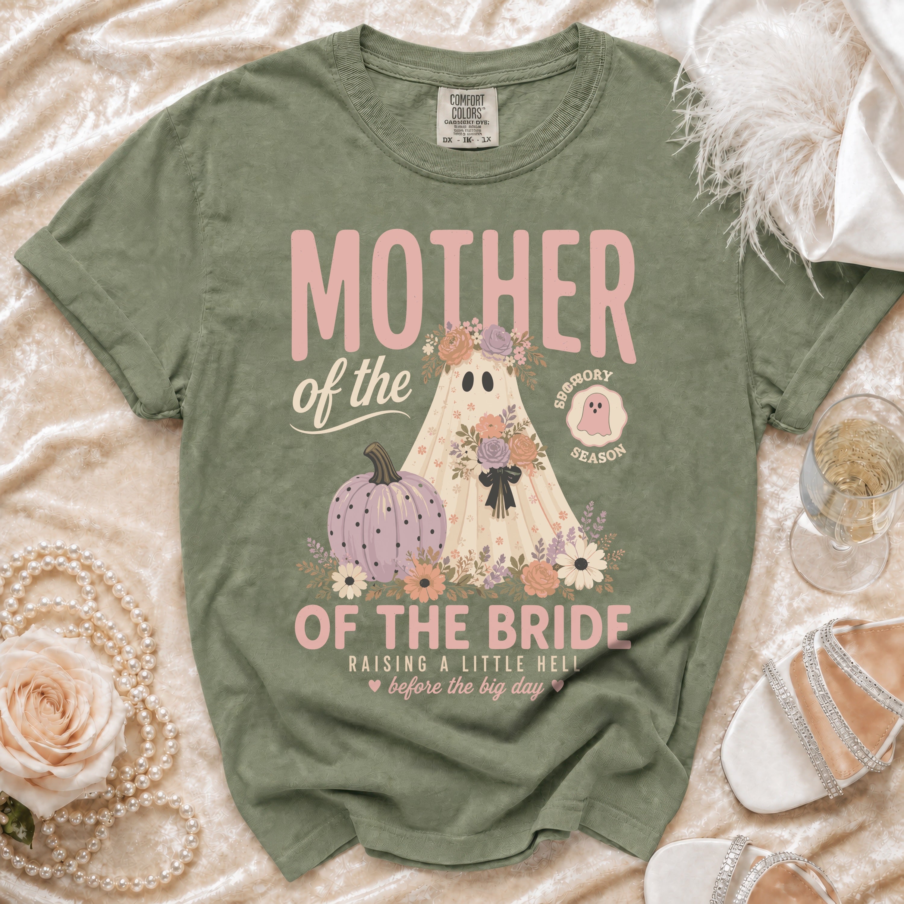 An olive green t-shirt with a vintage-style graphic of a ghost wearing floral wreaths and text reading Mother of the Bride.