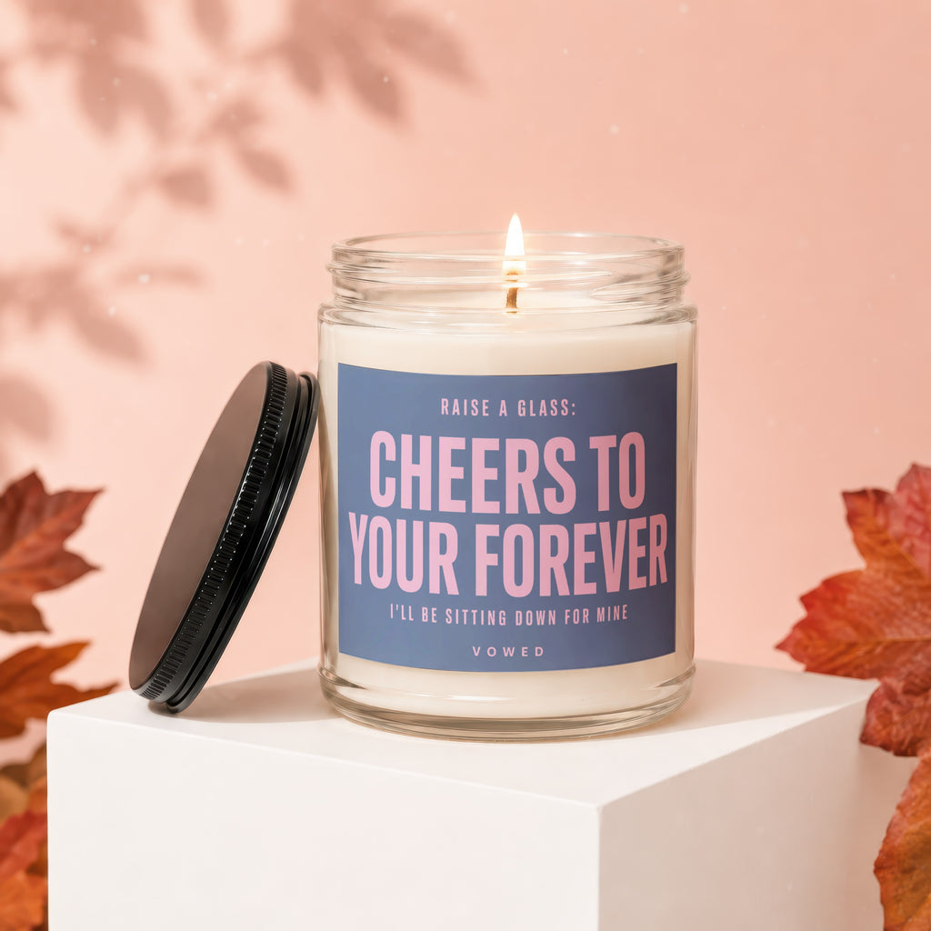 Glass candle with lit wick and lid off, labeled "Cheers to Your Forever".