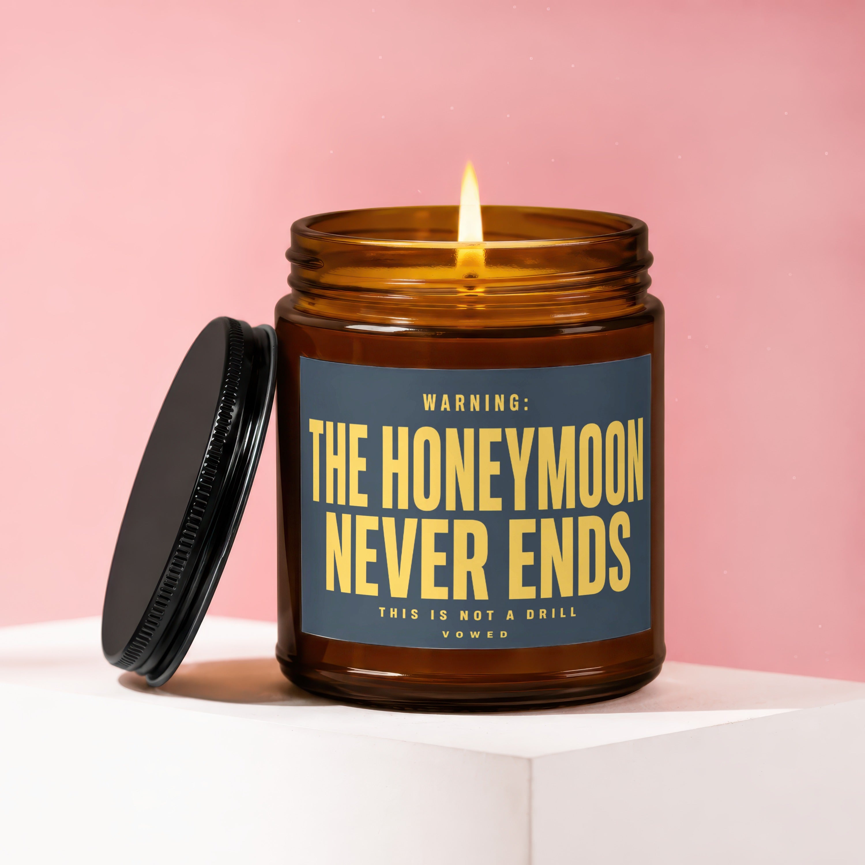 Amber-colored candle with lit flame, lid removed, reading "WARNING: THE HONEYMOON NEVER ENDS. THIS IS NOT A DRILL. VOWED"