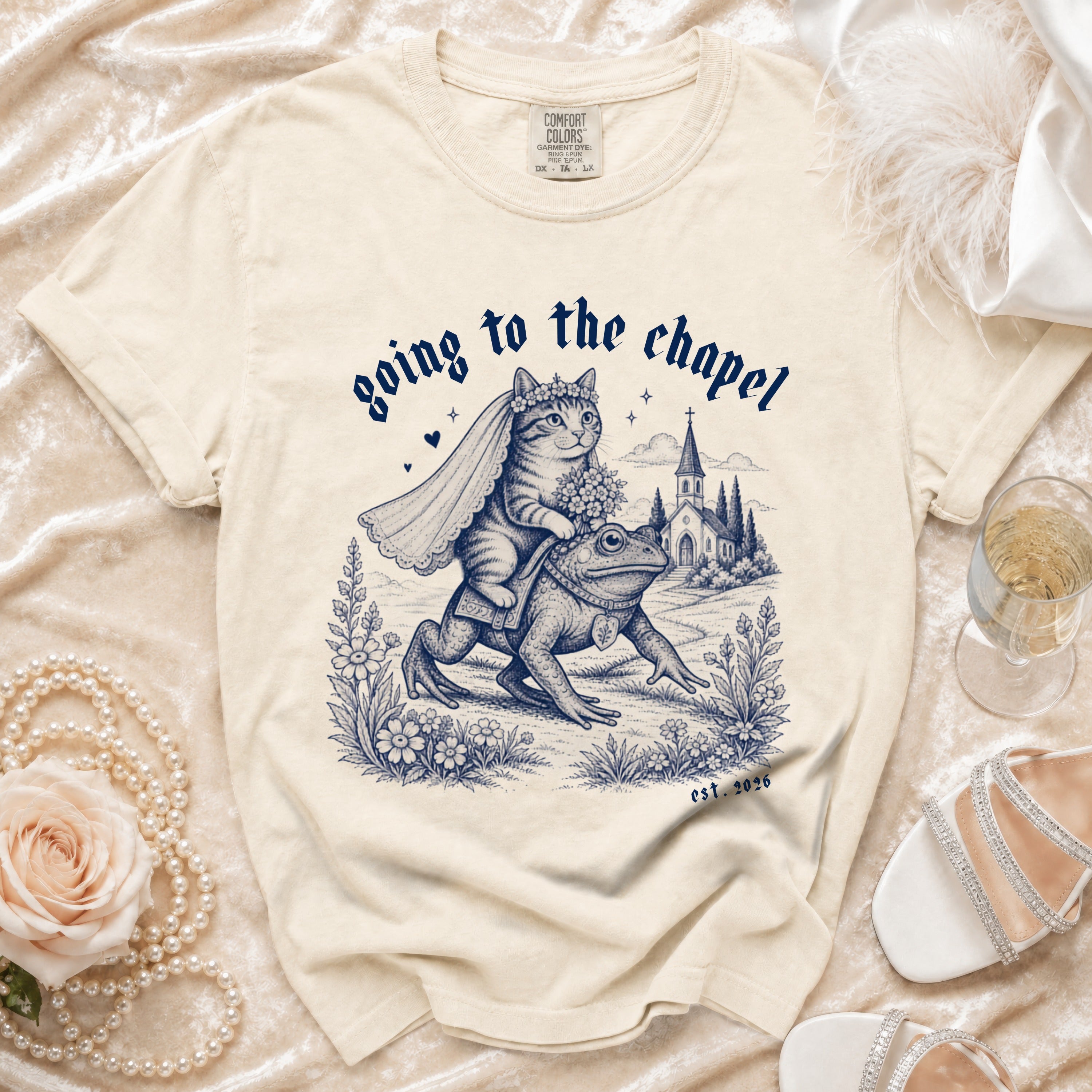Cream t-shirt with illustration of cat in bridal veil riding a frog to chapel, text "going to the chapel est. 2026".