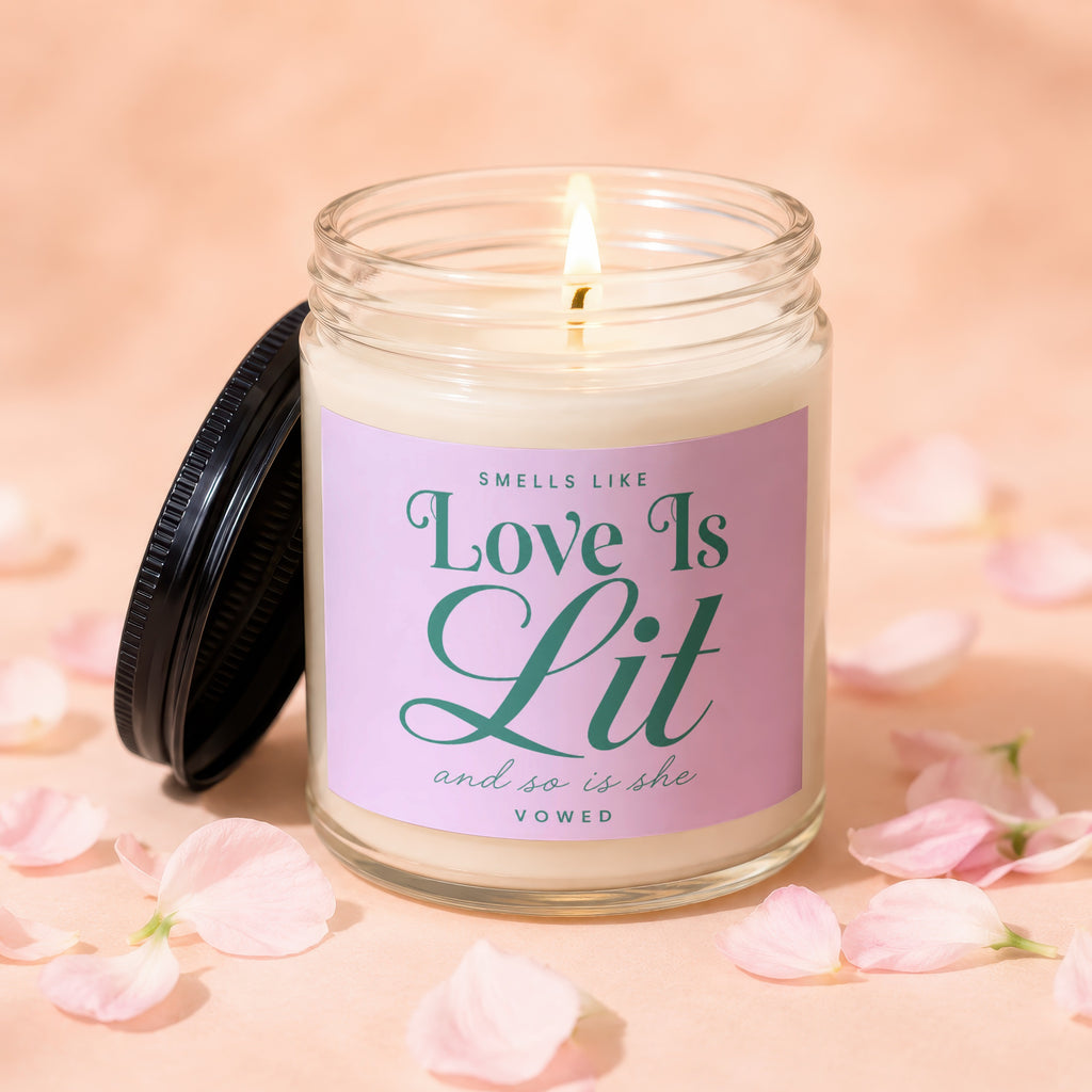 Glass candle, lit flame, with label that reads "Smells Like Love Is Lit and so is she VOWED."
