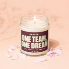 Glass jar candle with lit wick and burgundy label reading "ONE TEAM, ONE DREAM".