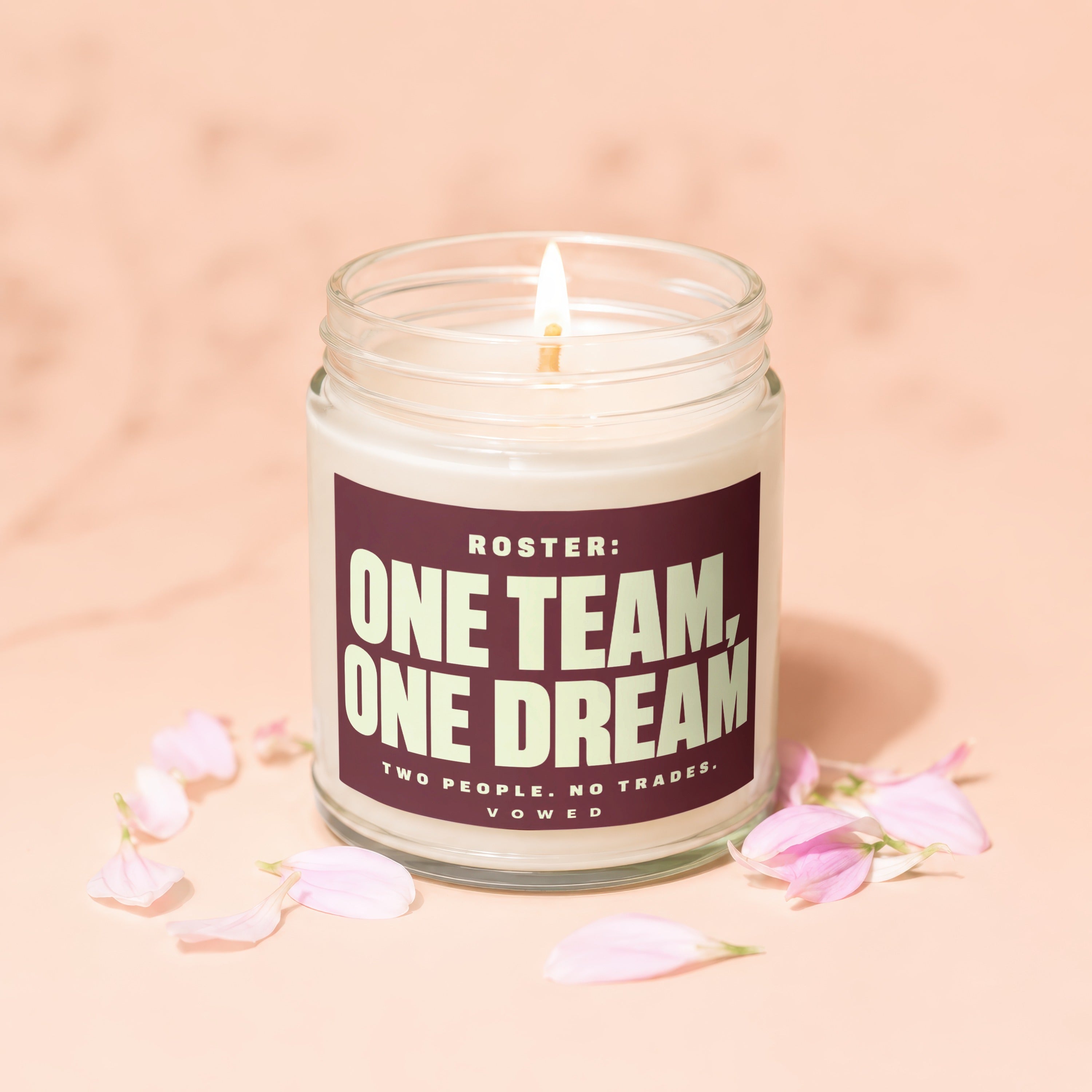 Glass jar candle with lit wick and burgundy label reading "ONE TEAM, ONE DREAM".