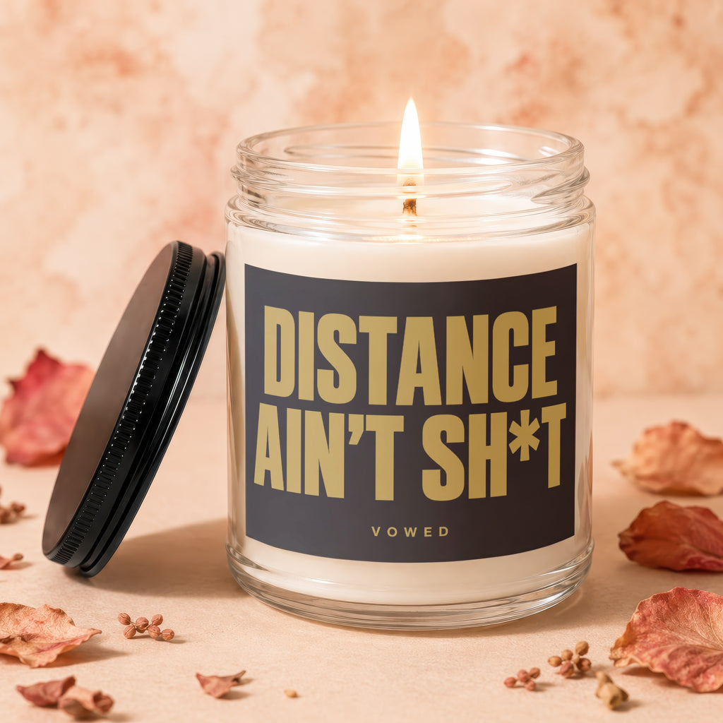Glass candle with burning wick, lid nearby, and label stating "DISTANCE AIN'T SH*T".