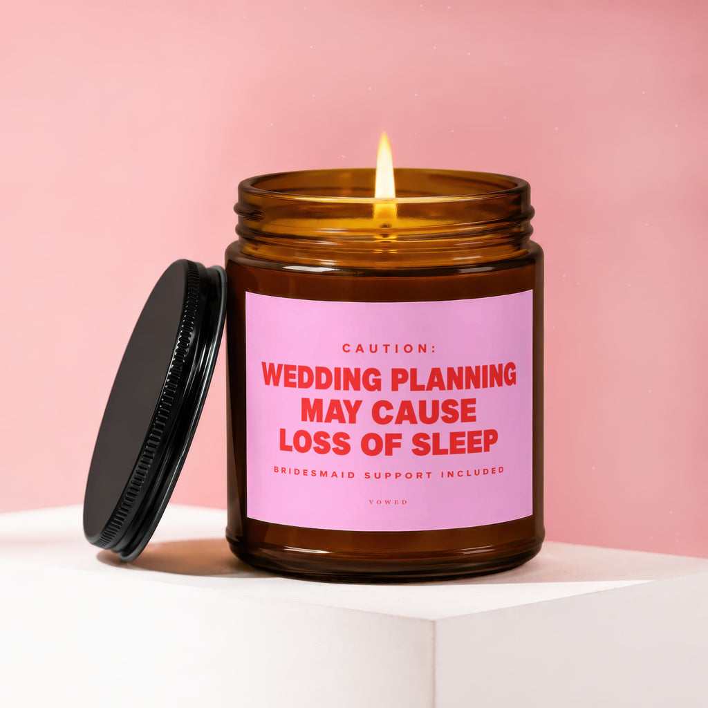 Amber glass candle with lit flame, pink label reads "Wedding planning may cause loss of sleep".