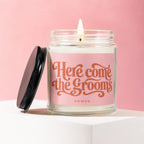 Candle with "Here Come the Grooms" text on pink label, lit flame, and black lid.