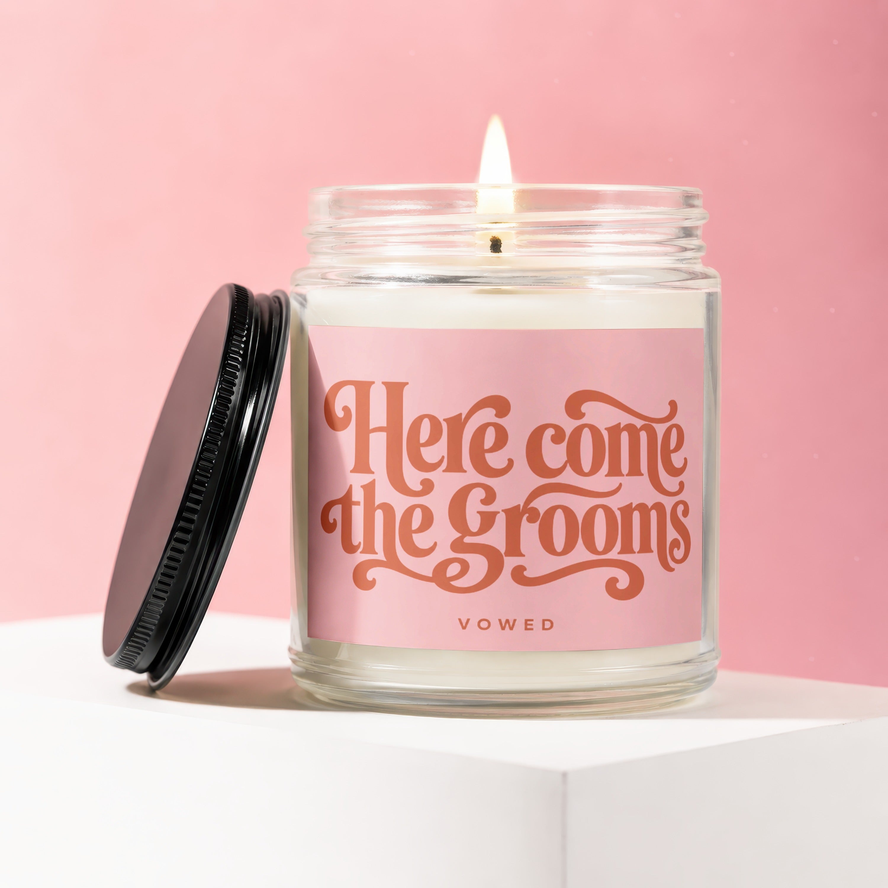 Candle with "Here Come the Grooms" text on pink label, lit flame, and black lid.