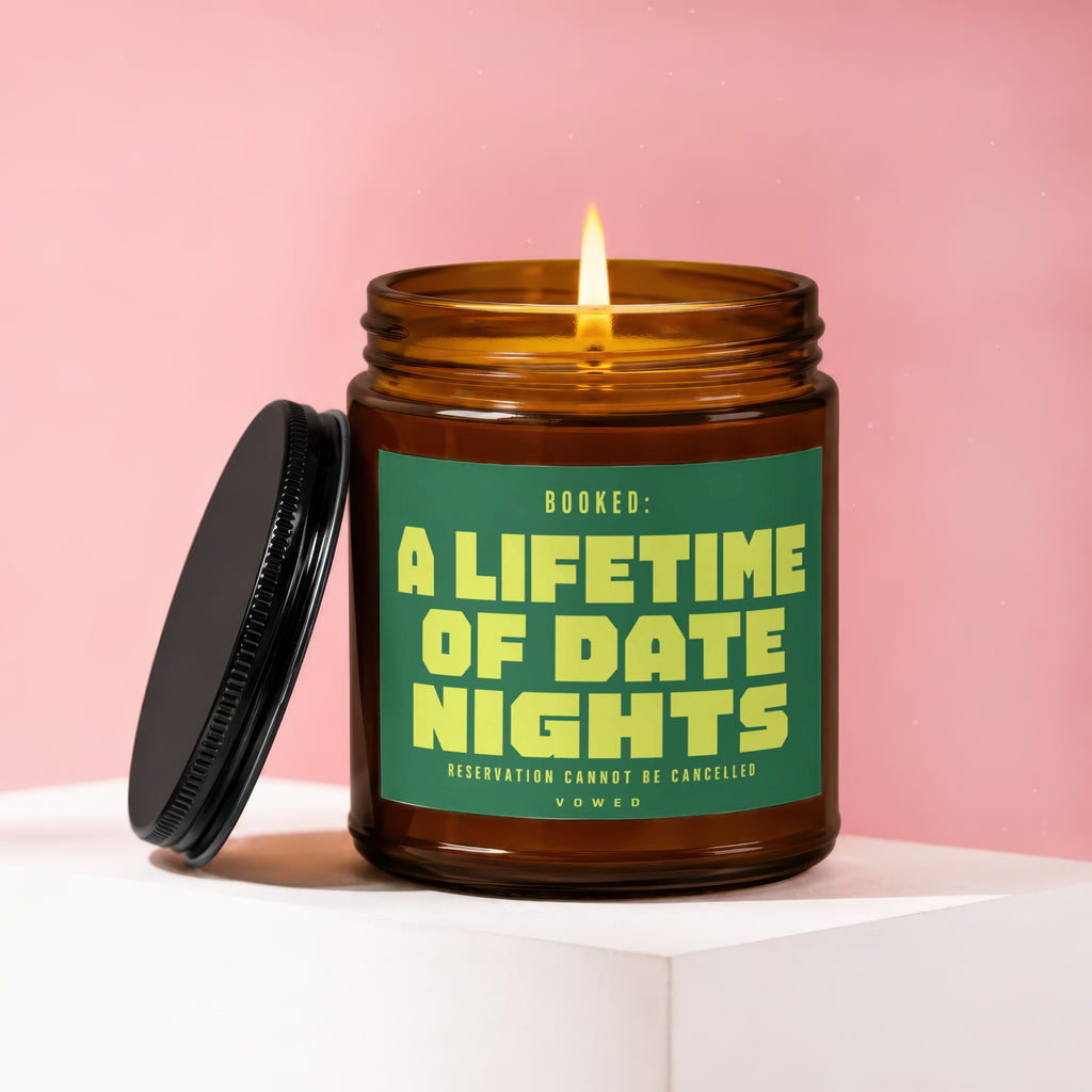 Amber jar candle with burning wick and black lid, reads "BOOKED: A LIFETIME OF DATE NIGHTS".