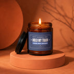 Amber jar candle with lit flame, navy label reads "Hold My Train And My Hand".