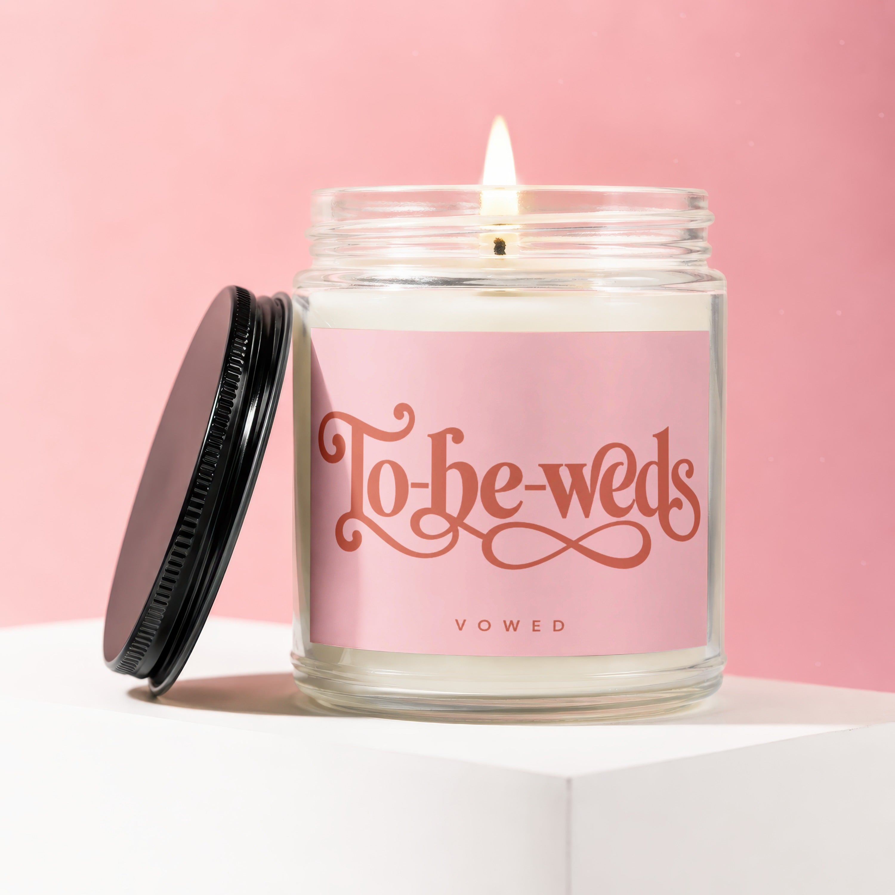 Glass candle with burning wick on pink background, says "To-be-weds" and "VOWED".