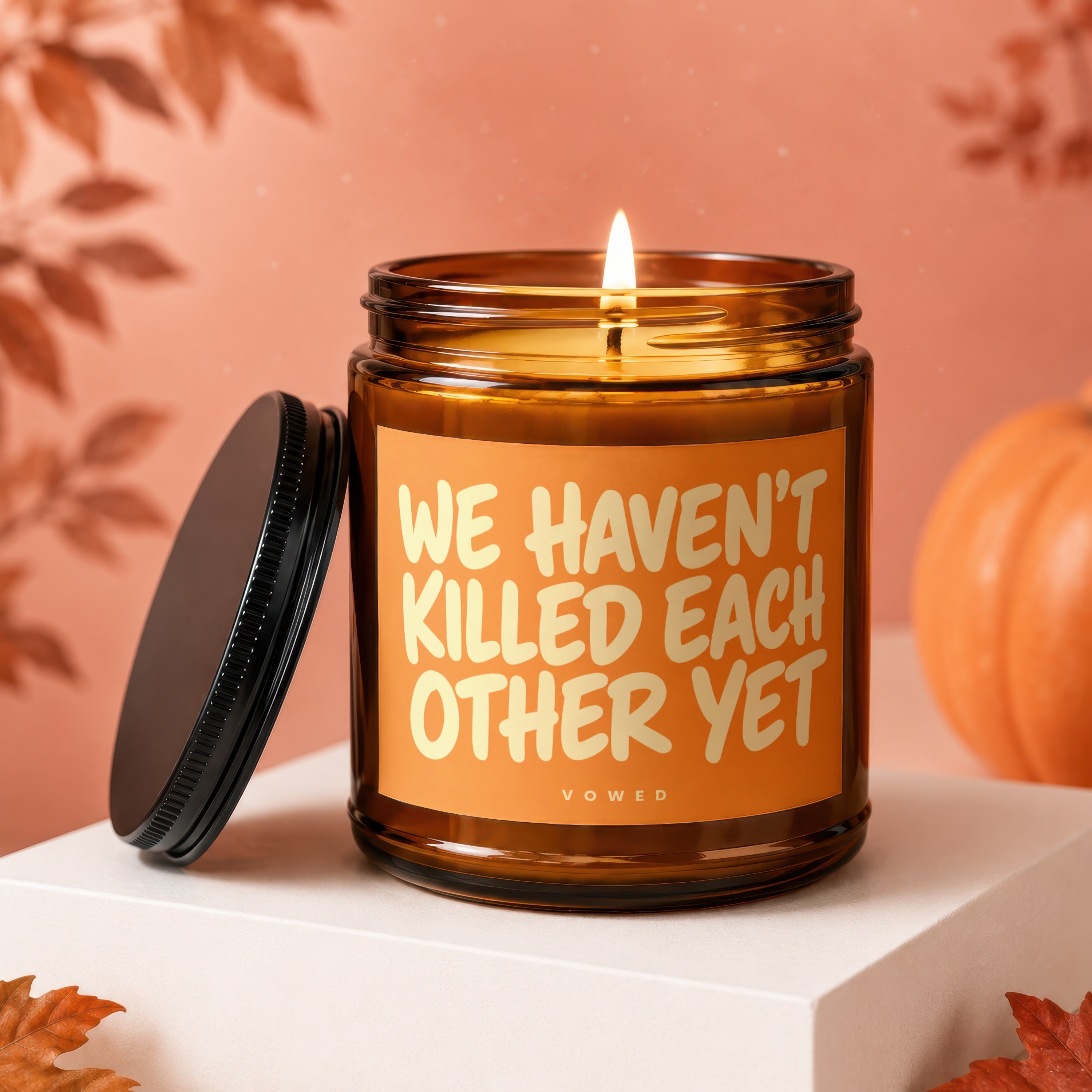Amber jar candle with lid off, lit flame, and "WE HAVEN'T KILLED EACH OTHER YET" text.