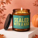 Amber jar candle with "SEALED WITH A KISS" text, lit flame, pumpkin, and autumn leaves.