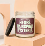 Glass jar candle with dark purple label reading "Ingredients: Hexes. Hairspray. Hysteria."