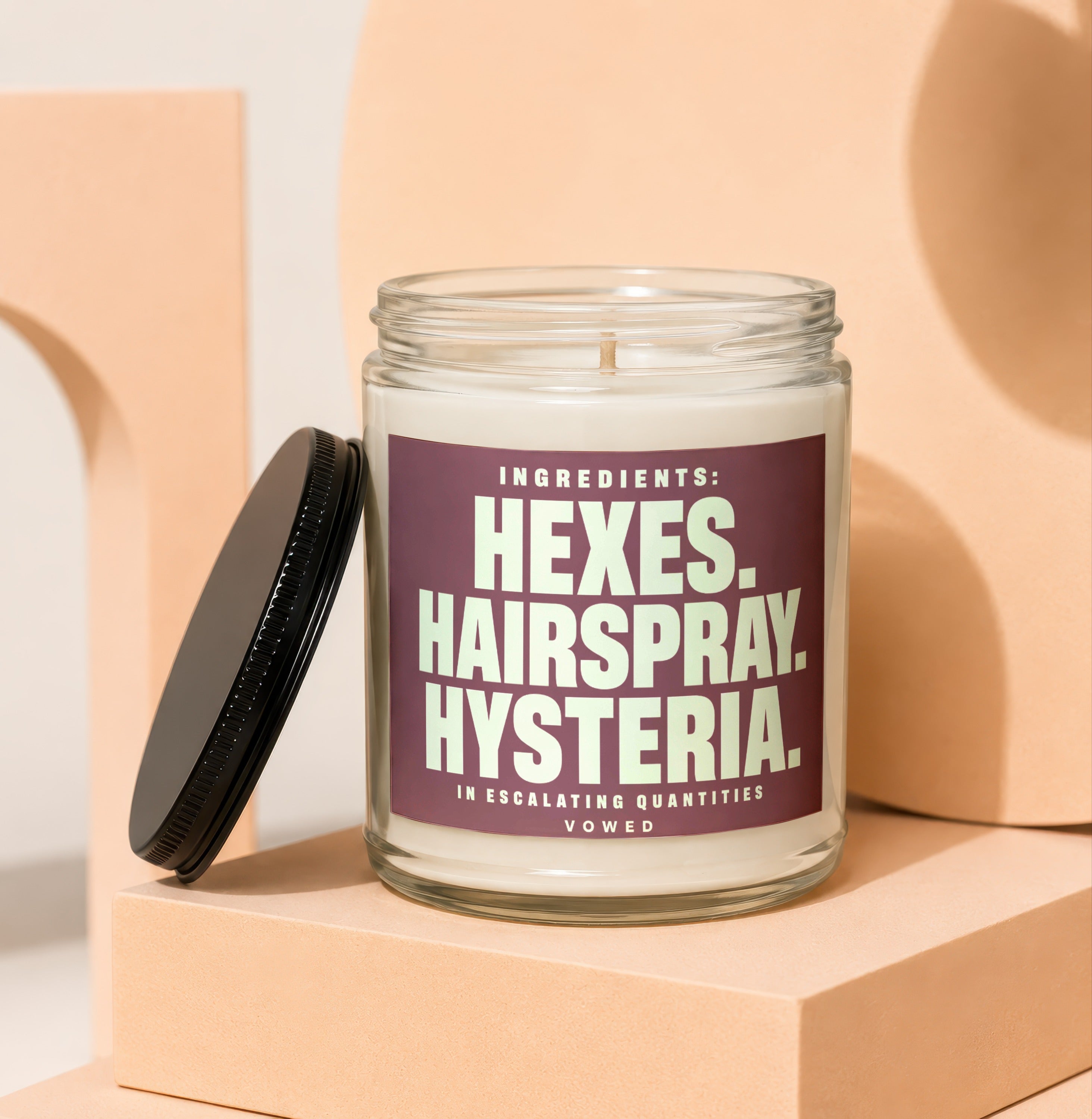 Glass jar candle with dark purple label reading "Ingredients: Hexes. Hairspray. Hysteria."