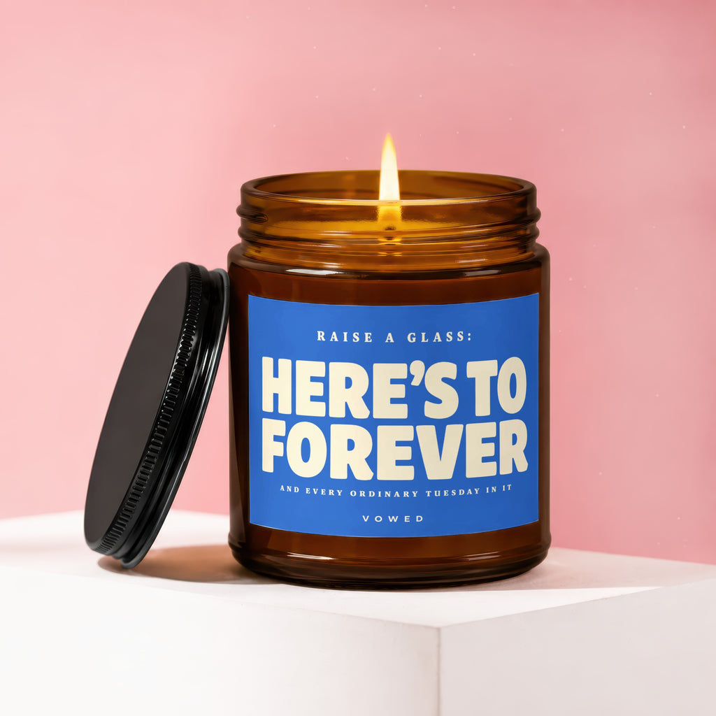 Amber glass candle with lit wick and black lid, featuring blue label with text: "RAISE A GLASS: HERE'S TO FOREVER"
