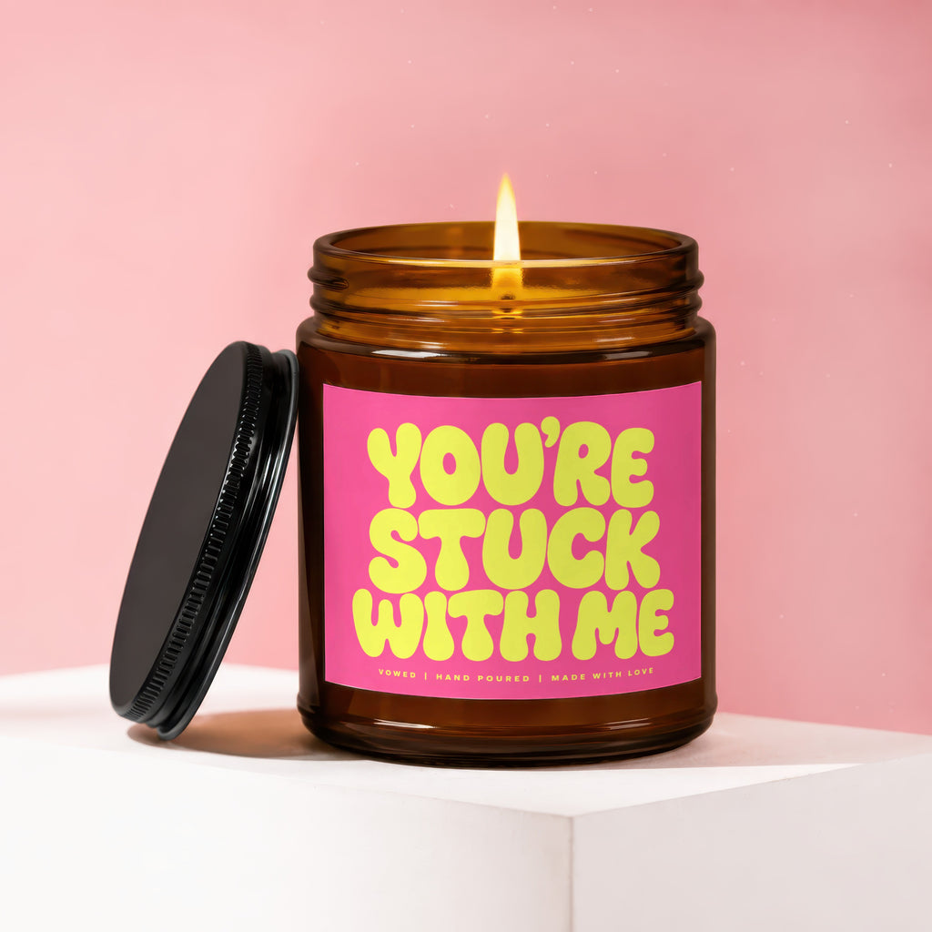 Amber jar candle with pink label reading "YOU'RE STUCK WITH ME" and a lit wick.
