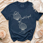 A navy blue t-shirt with a white graphic of two seashells tied together, a cowboy hat on top, and the text "Coastal Cowgirl BACH EST. 2026."