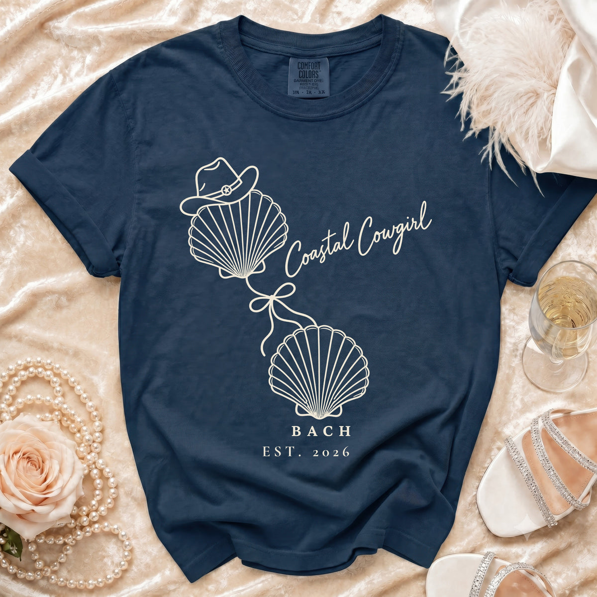 A navy blue t-shirt with a white graphic of two seashells tied together, a cowboy hat on top, and the text "Coastal Cowgirl BACH EST. 2026."