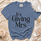 Blue t-shirt with "It's Giving Mrs" text, styled with pearls, rose, and champagne.