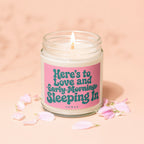 Burning candle in jar with pink label reading "Here's to Love and Early Mornings Sleeping In".