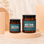 Two amber glass candles with black lids, featuring labels that read "Saved for you: Next to Me Maid of Honor Spot".